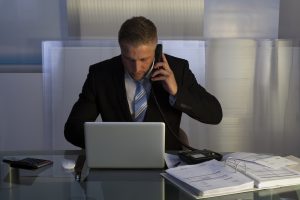 Stressed businessman working late into the night putting in overtime to meet a deadline sitting at his desk using a laptop and talking on his mobile