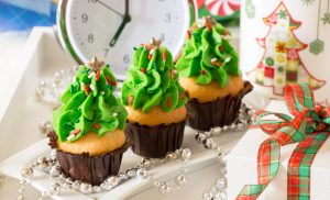 Christmas cupcakes mug of tea gift holiday decorated on white tray behind alarm clock. Christmas trees shape capcakes for festive breakfast. Horizontal. Close.